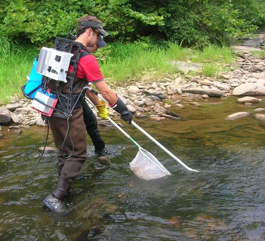 Crew members wade through a stream, carrying equipment.